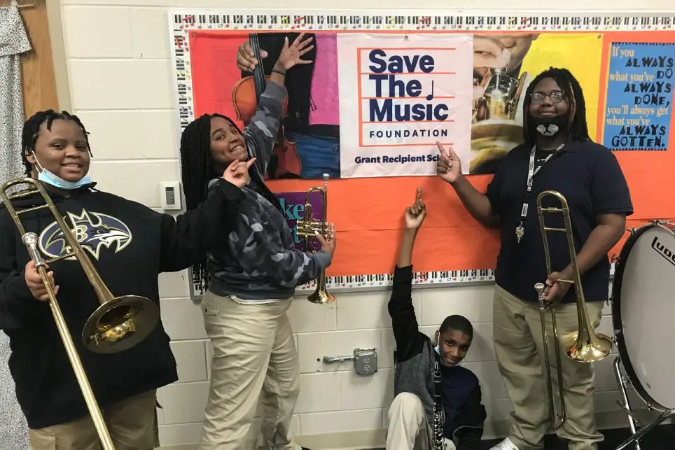 A group of students holding their instruments and pointing at a Save The Music Foundation poster.